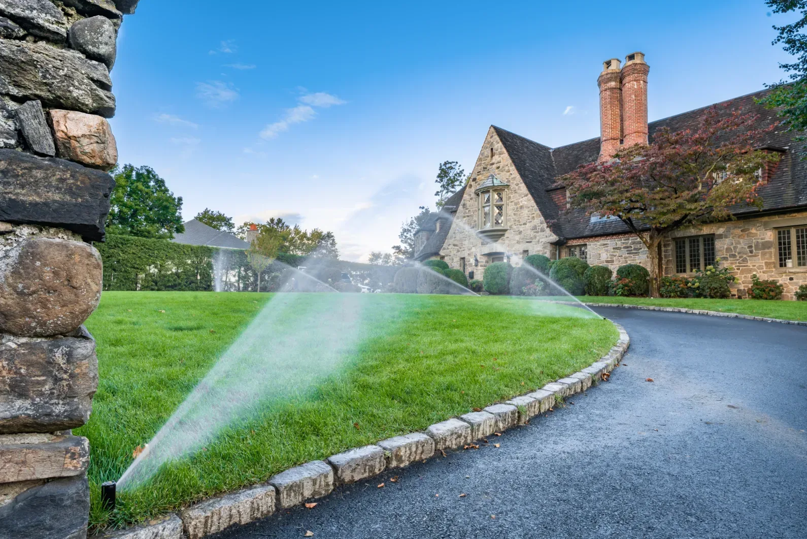 Pop-up sprinklers spraying the lawn beside a curved driveway and stone house.