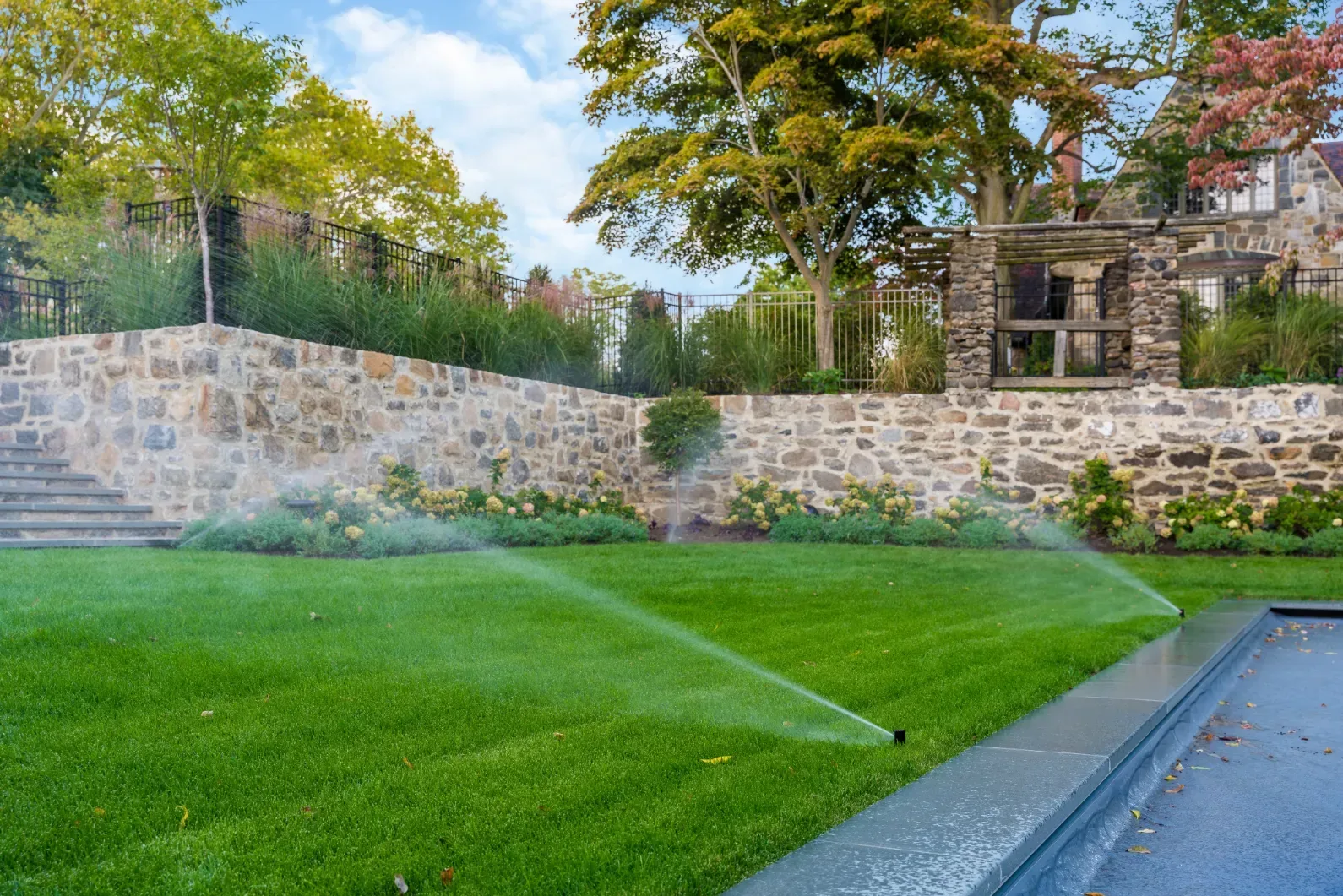 Sprinklers watering a lawn below a stone retaining wall beside a pool.