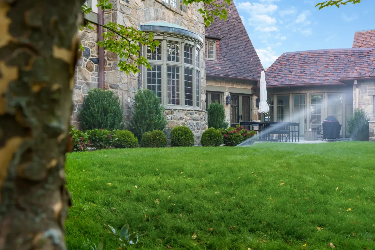 Sprinkler spray across a small lawn beside a stone house.