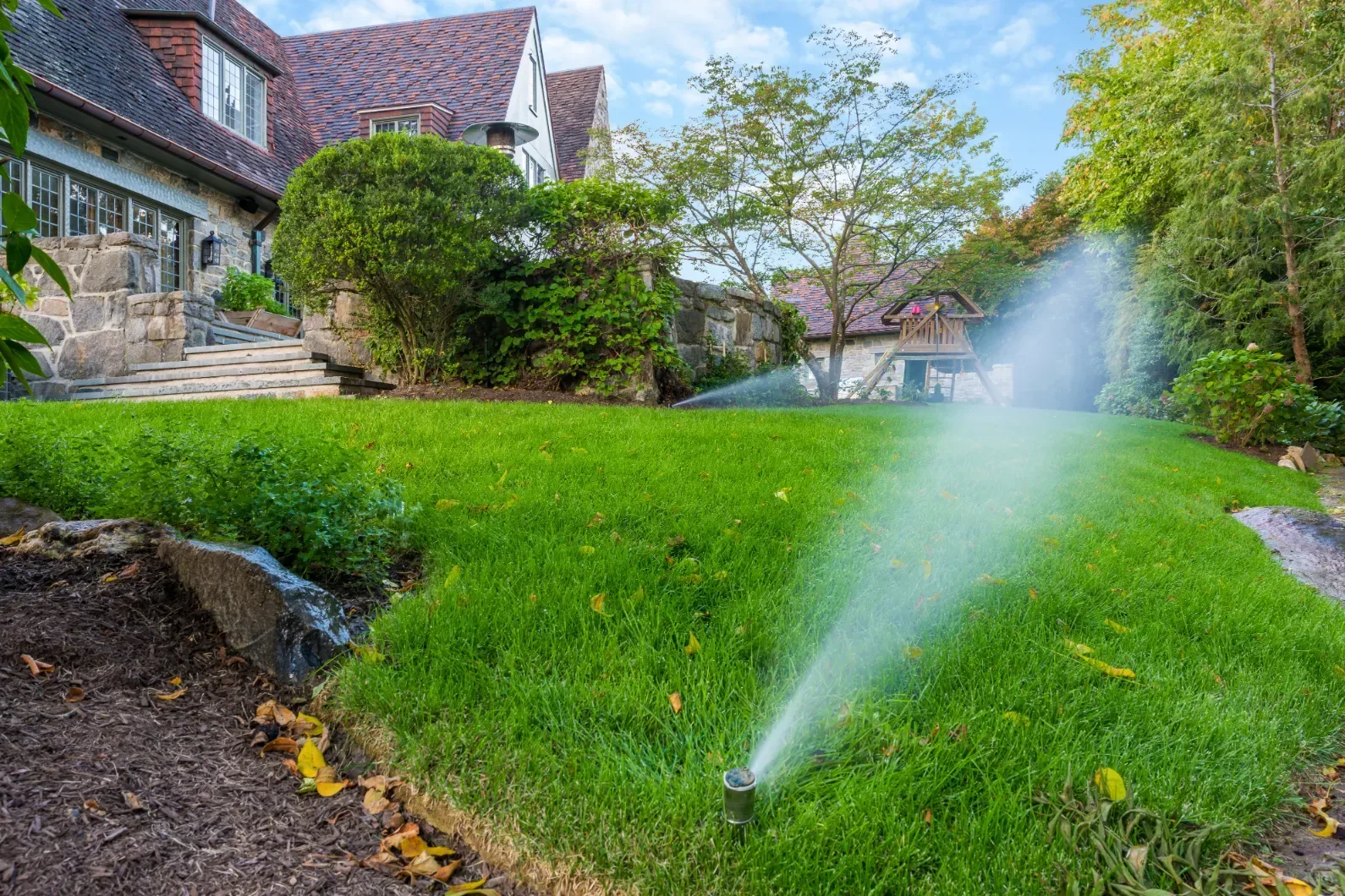 Sprinkler spray across a small lawn beside a stone house.