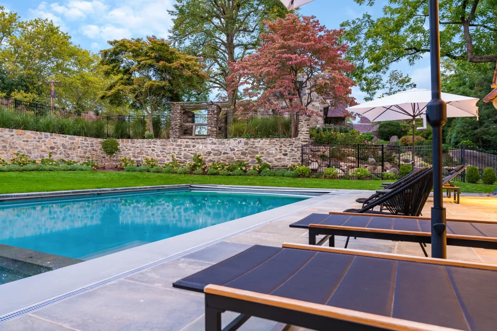 Poolside lounge chairs facing a stone retaining wall and trees in daylight.