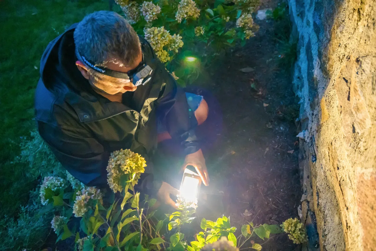 A worker tending a landscape light beside a stone wall after dark.