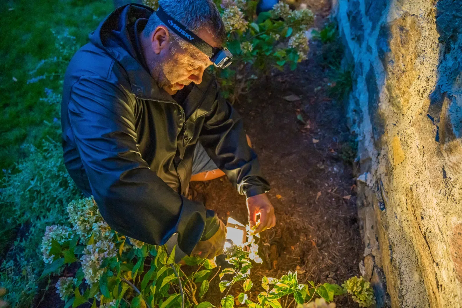 A worker tending a landscape light beside a stone wall after dark.