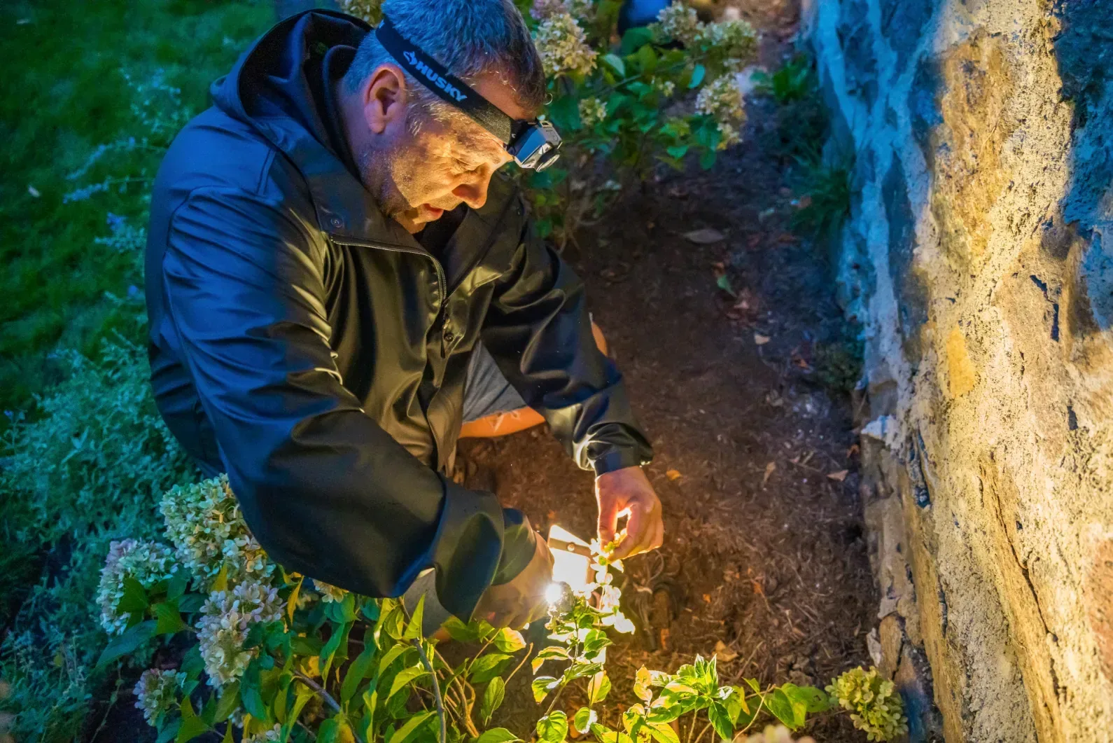 A worker tending a landscape light beside a stone wall after dark.