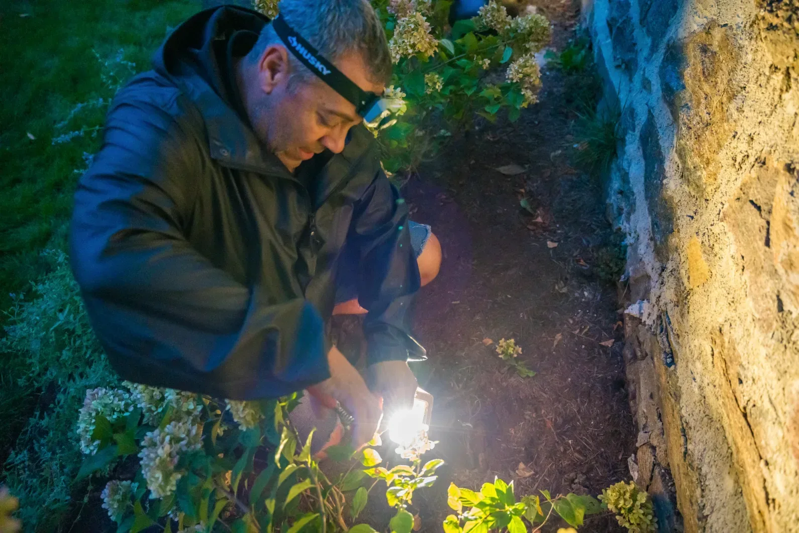 A worker tending a landscape light beside a stone wall after dark.