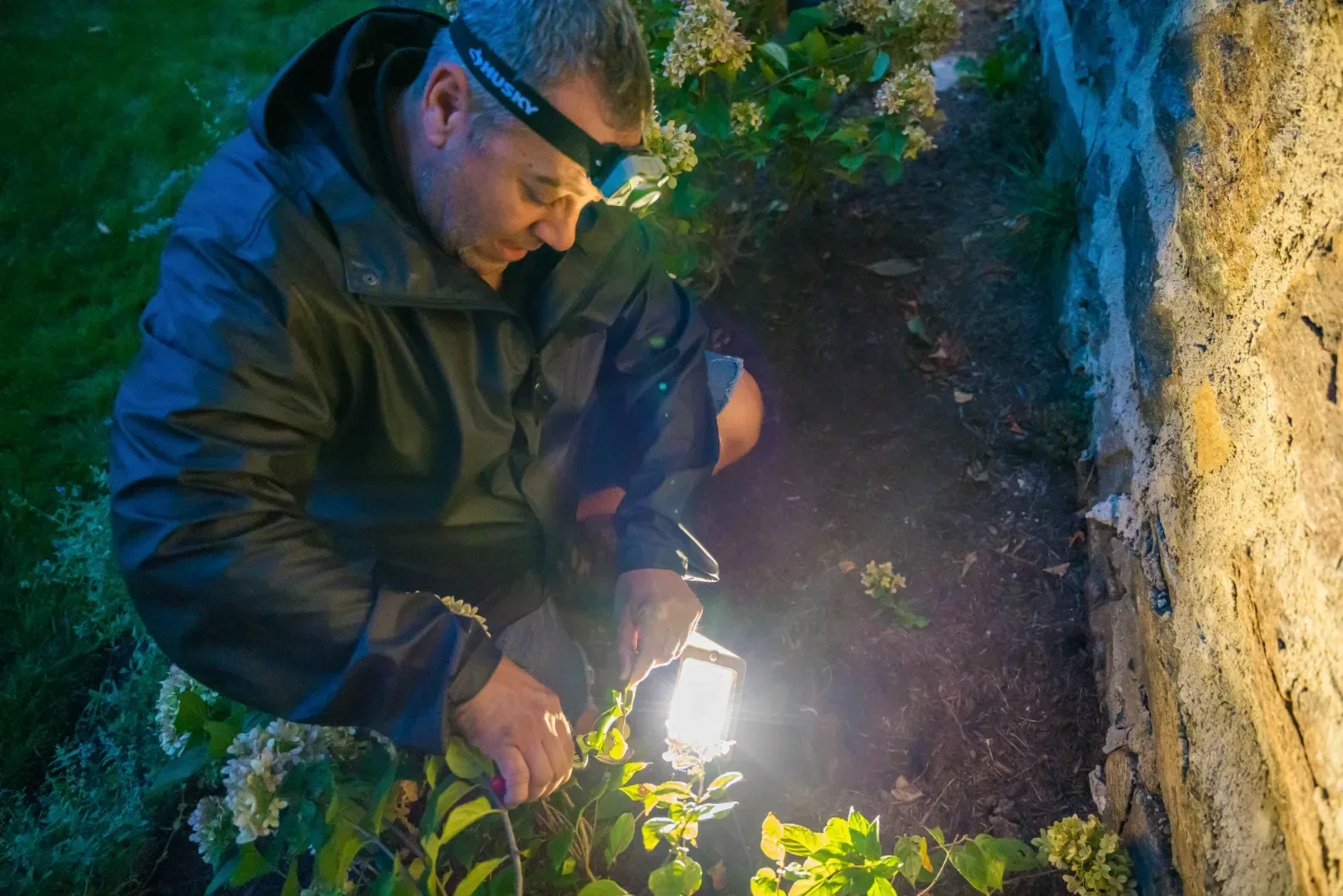 A worker tending a landscape light beside a stone wall after dark.