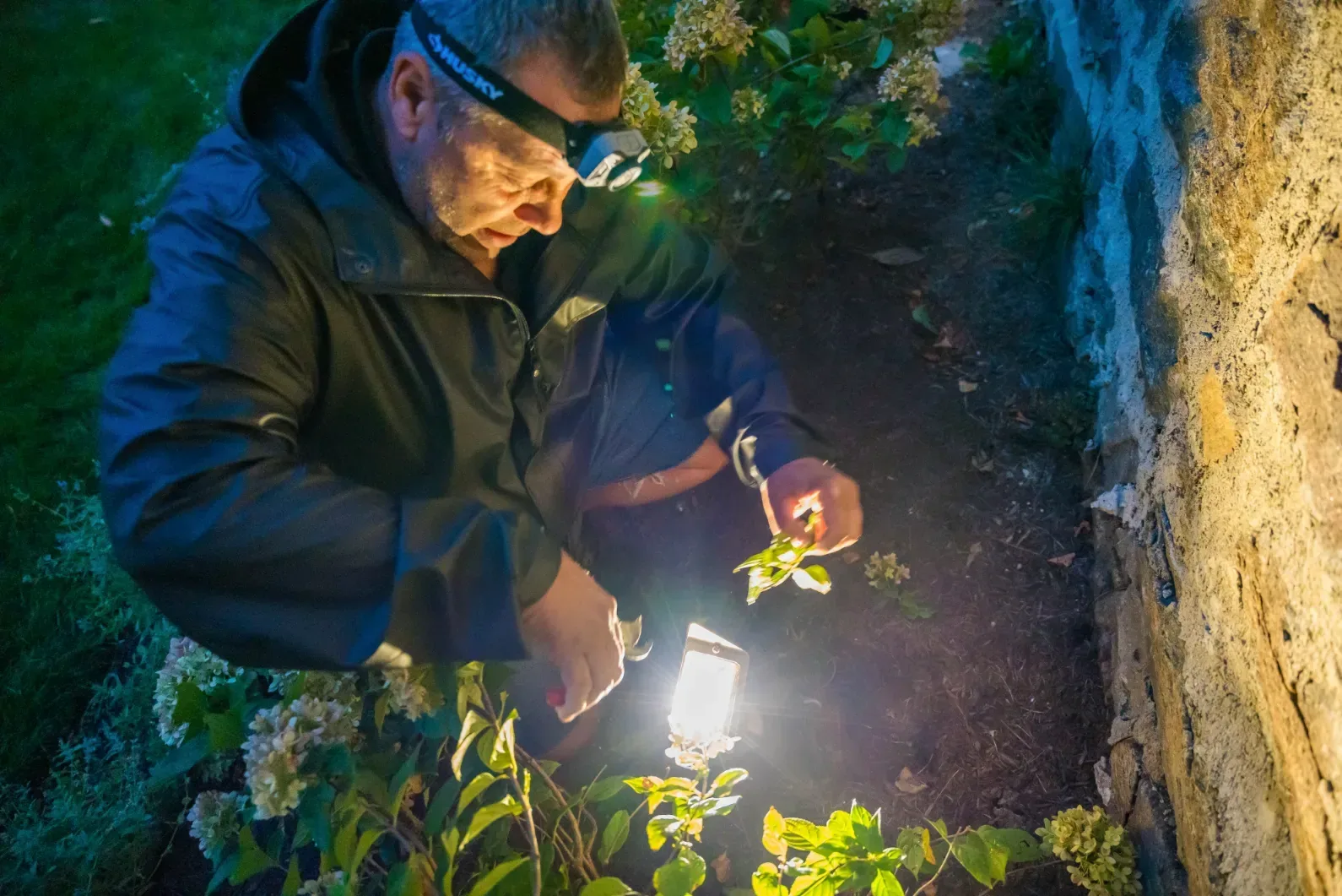 A worker tending a landscape light beside a stone wall after dark.