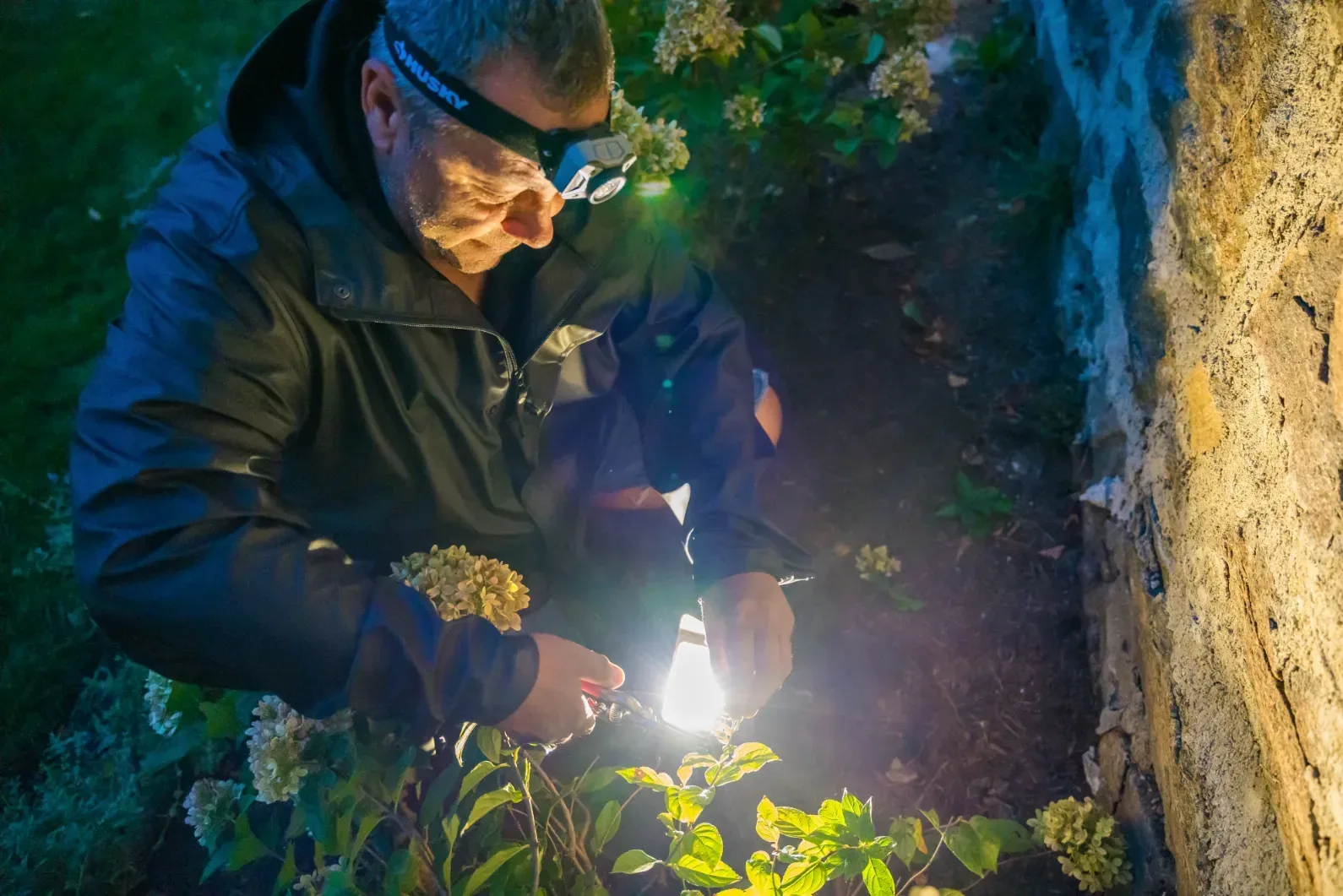 A worker tending a landscape light beside a stone wall after dark.