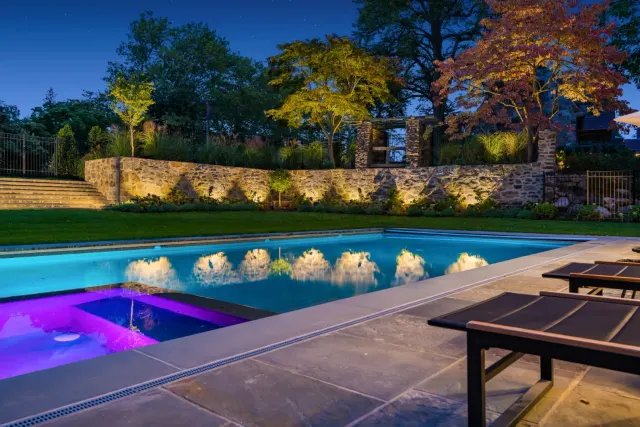 Swimming pool and spa reflecting a lit stone wall and trees after dark.
