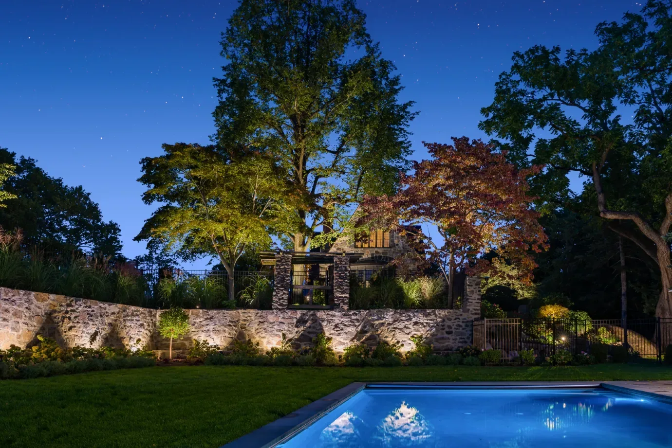 Illuminated stone retaining wall and trees above a blue-lit swimming pool.