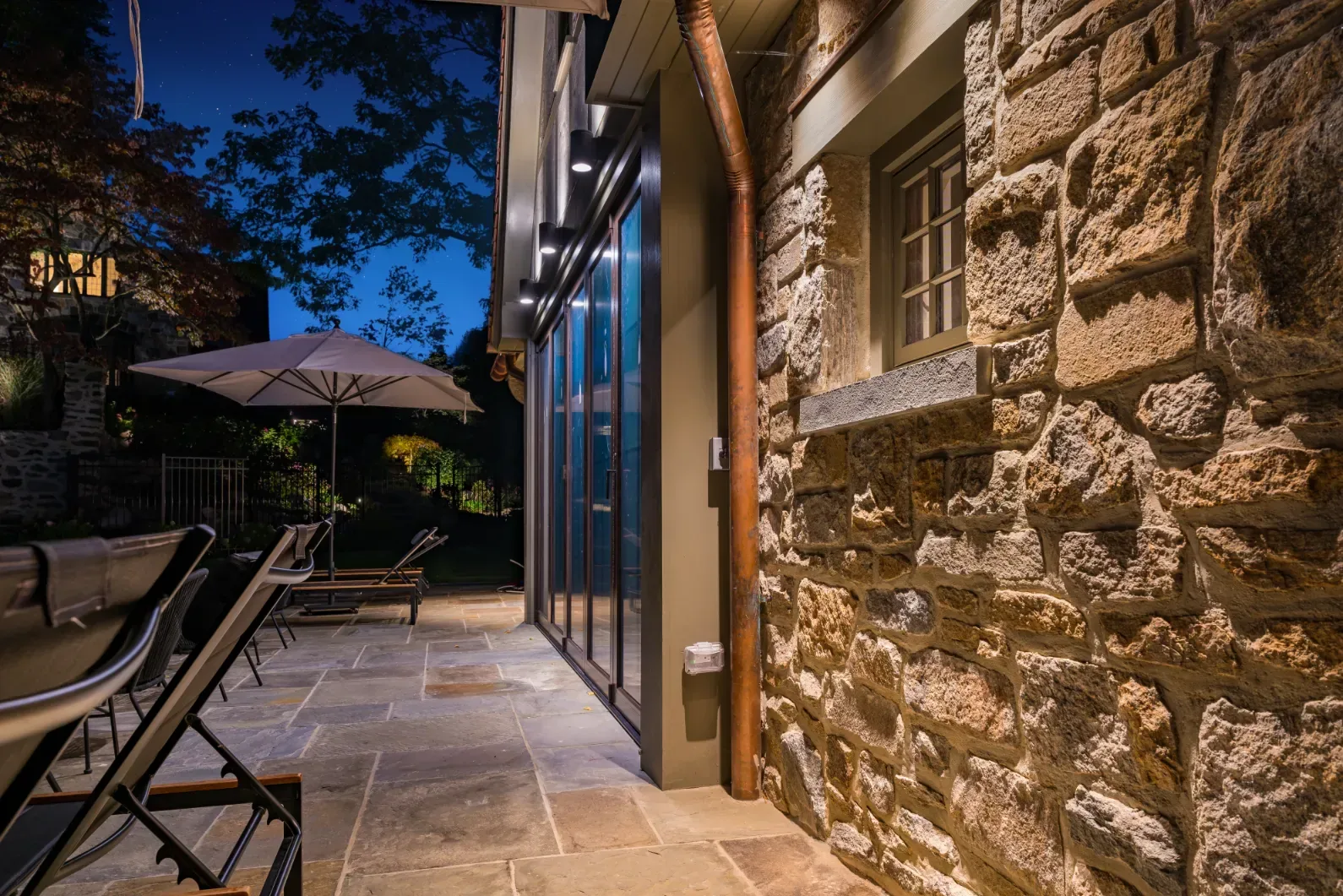 Textured stone pool-house wall illuminated beside a paved terrace.