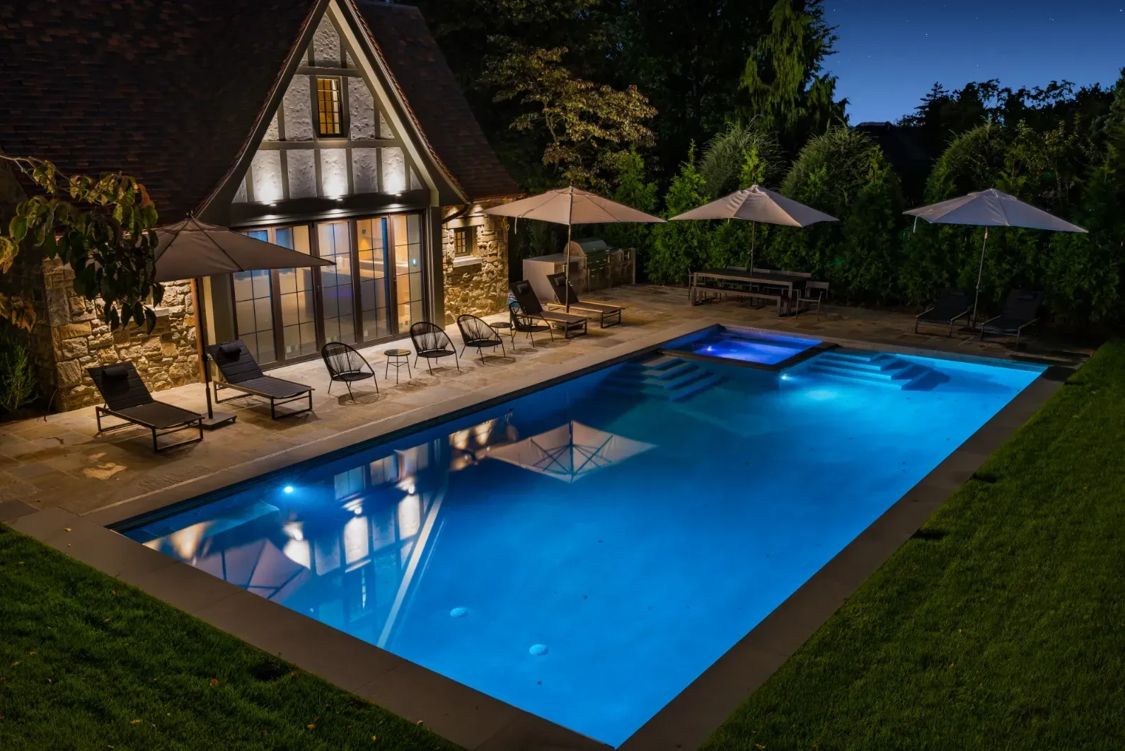 Blue-lit swimming pool beside an illuminated pool house and patio umbrellas.