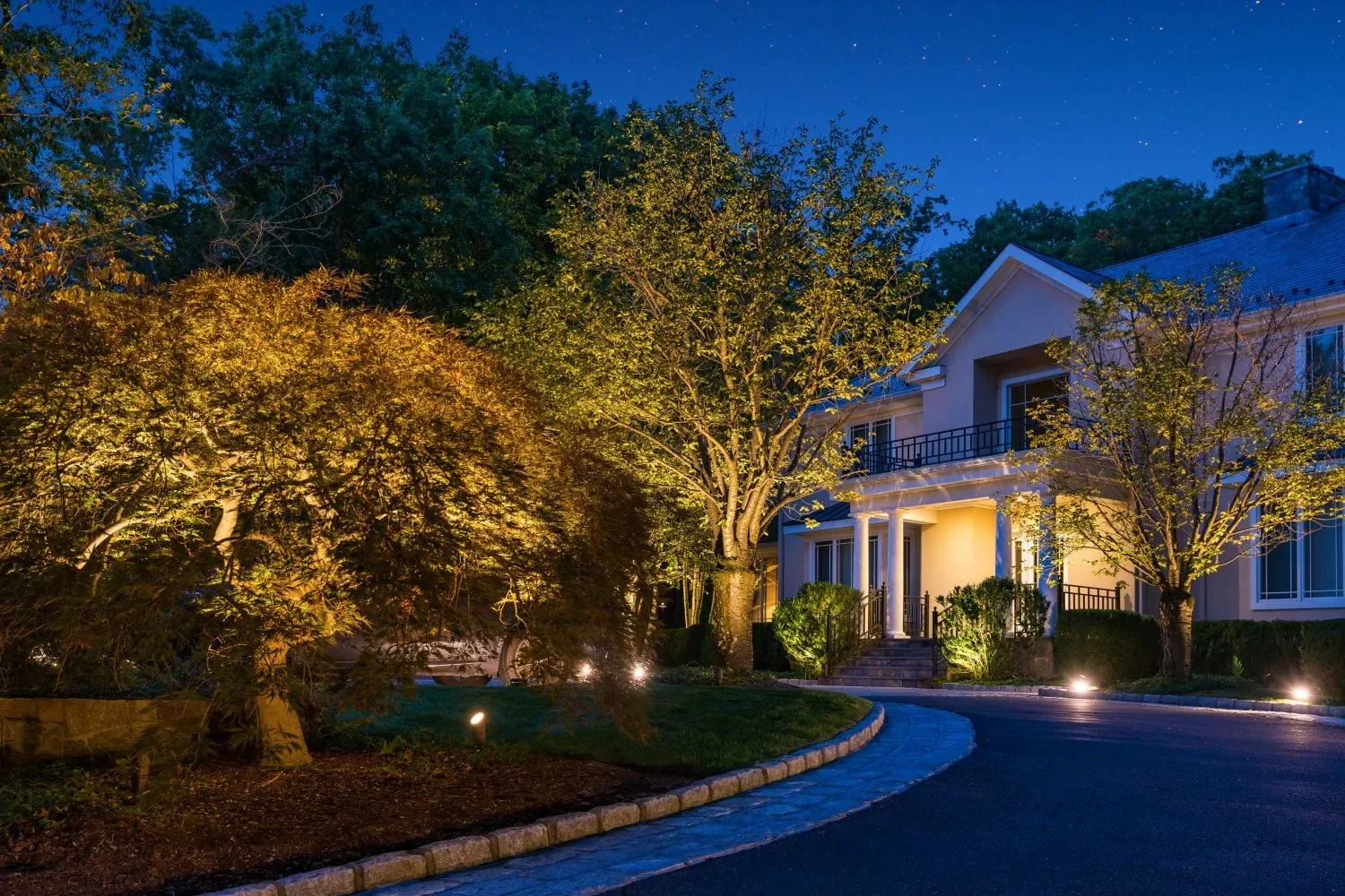 Illuminated house, trees, and planting beds along a curved driveway.