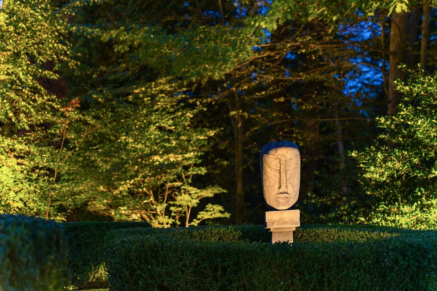 Sculpted stone face illuminated above clipped hedges in a garden.