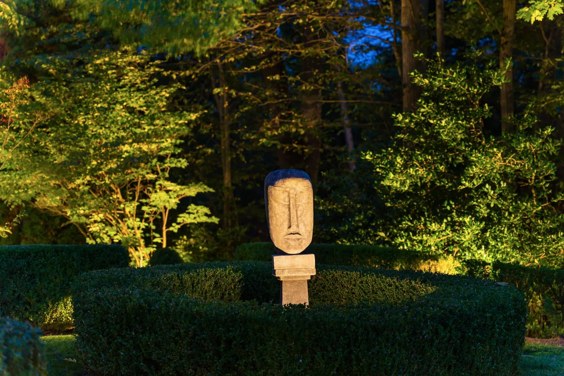 Sculpted stone face illuminated above clipped hedges in a garden.