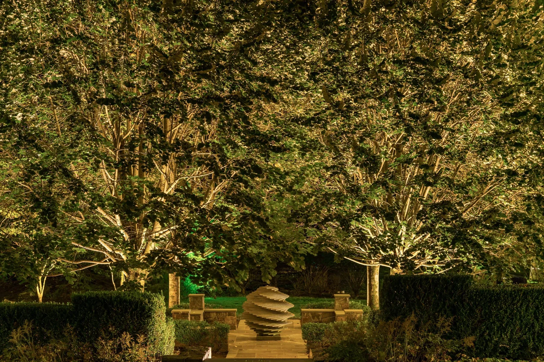 Two illuminated trees framing a stone sculpture within a formal garden.