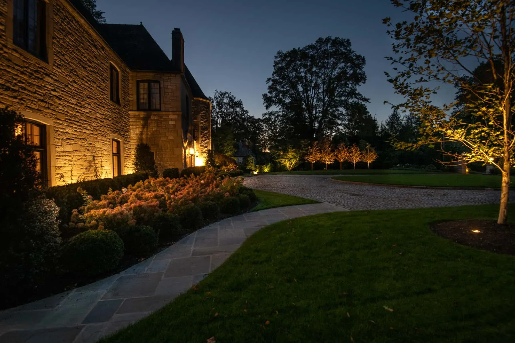 Lighting on a stone house beside a walkway, lawn, and ornamental trees.