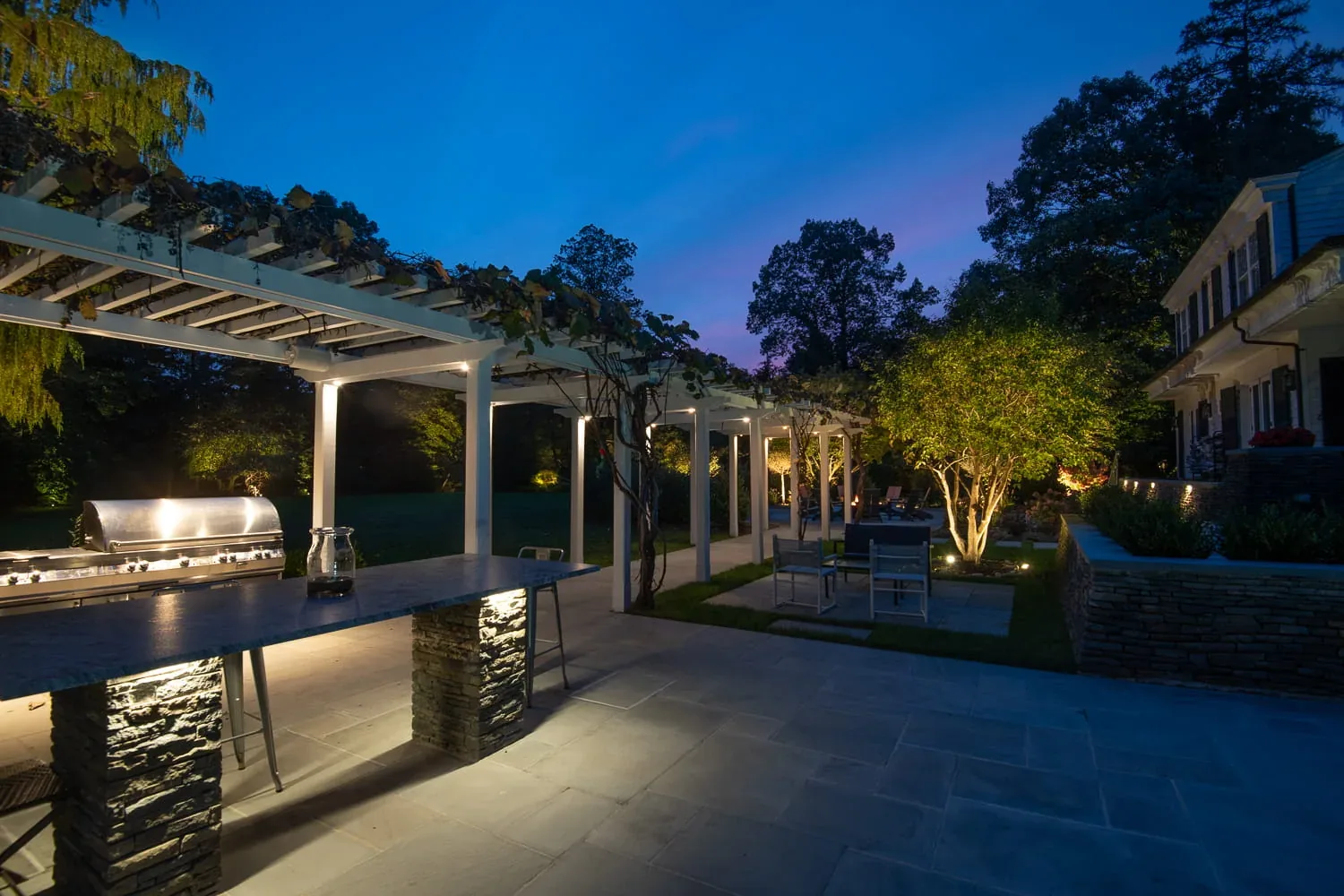 Outdoor kitchen and long pergola illuminated beside a garden terrace at dusk.