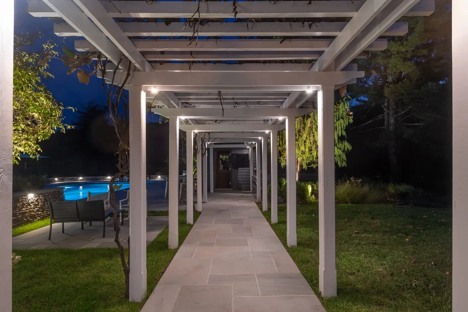 Lights along a white pergola above a stone path beside a pool at night.