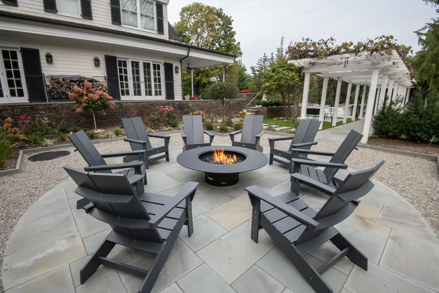 Adirondack chairs around a burning fire pit beside a white pergola.
