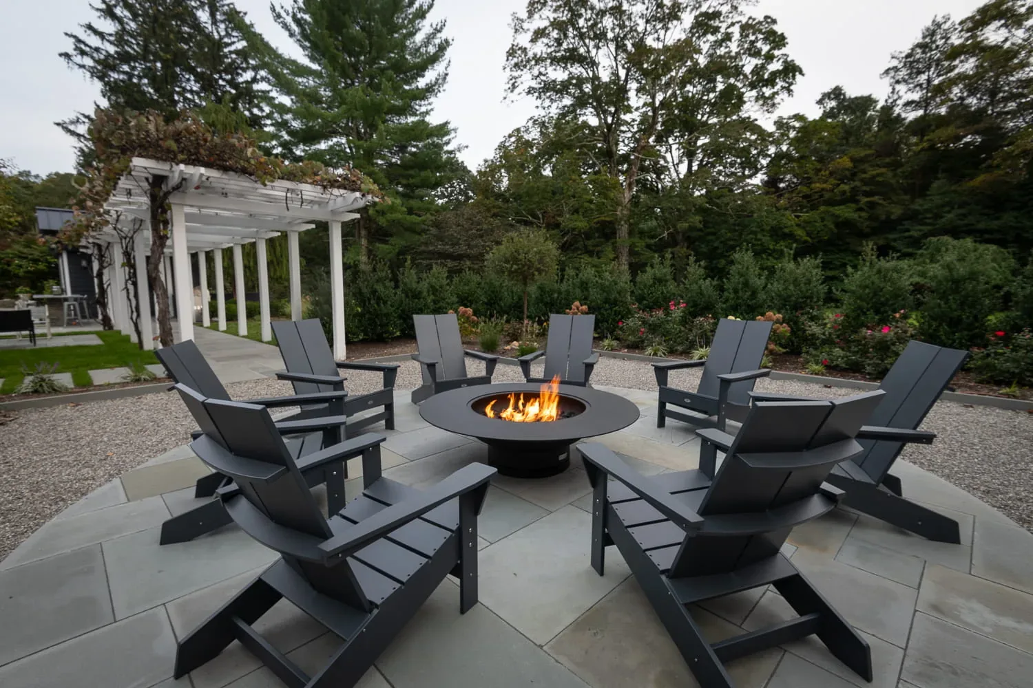Adirondack chairs around a burning fire pit beside a white pergola.