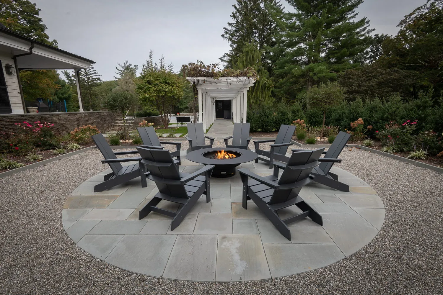 Adirondack chairs around a burning fire pit beside a white pergola.