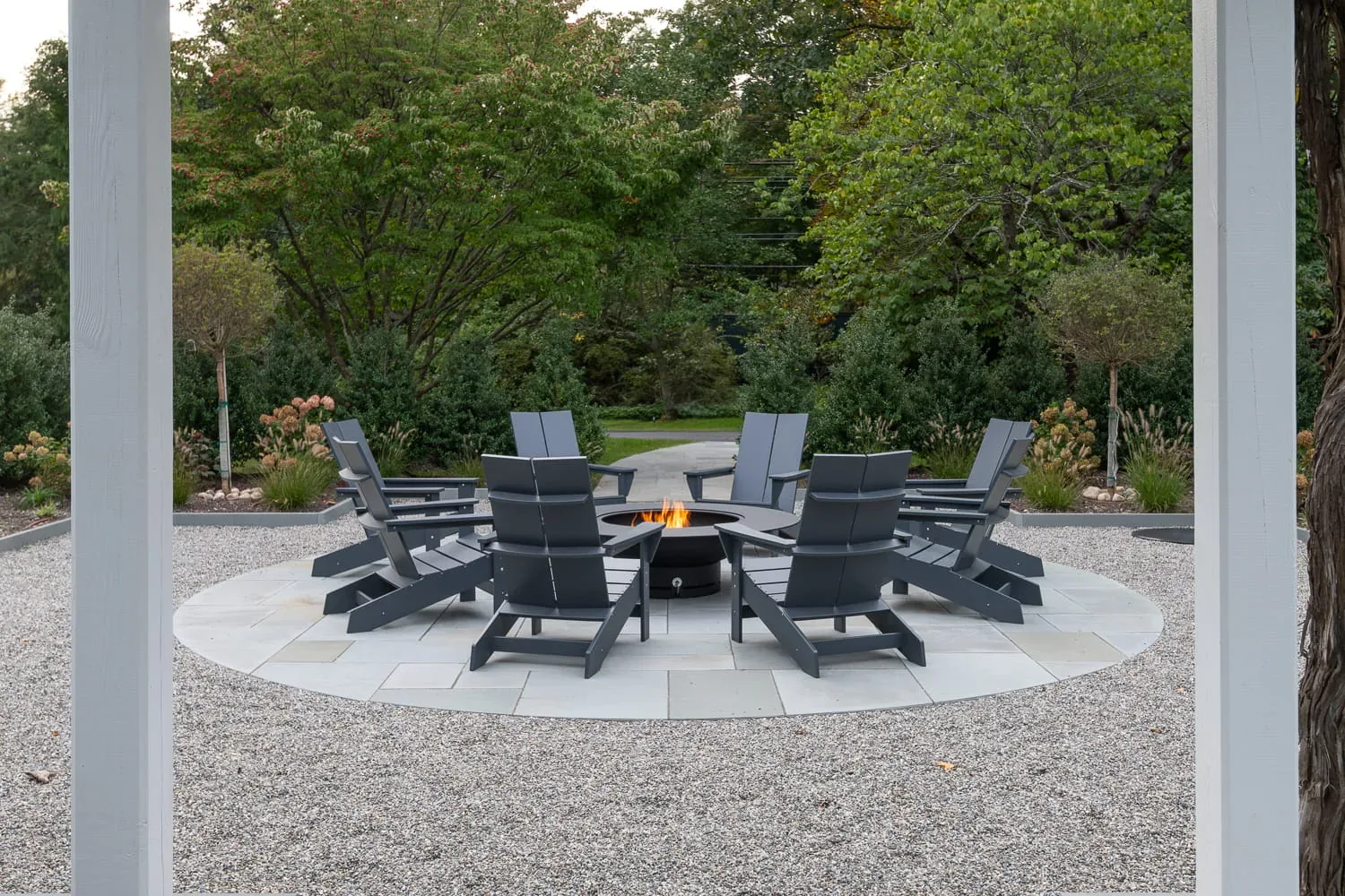 Adirondack chairs around a burning fire pit beside a white pergola.