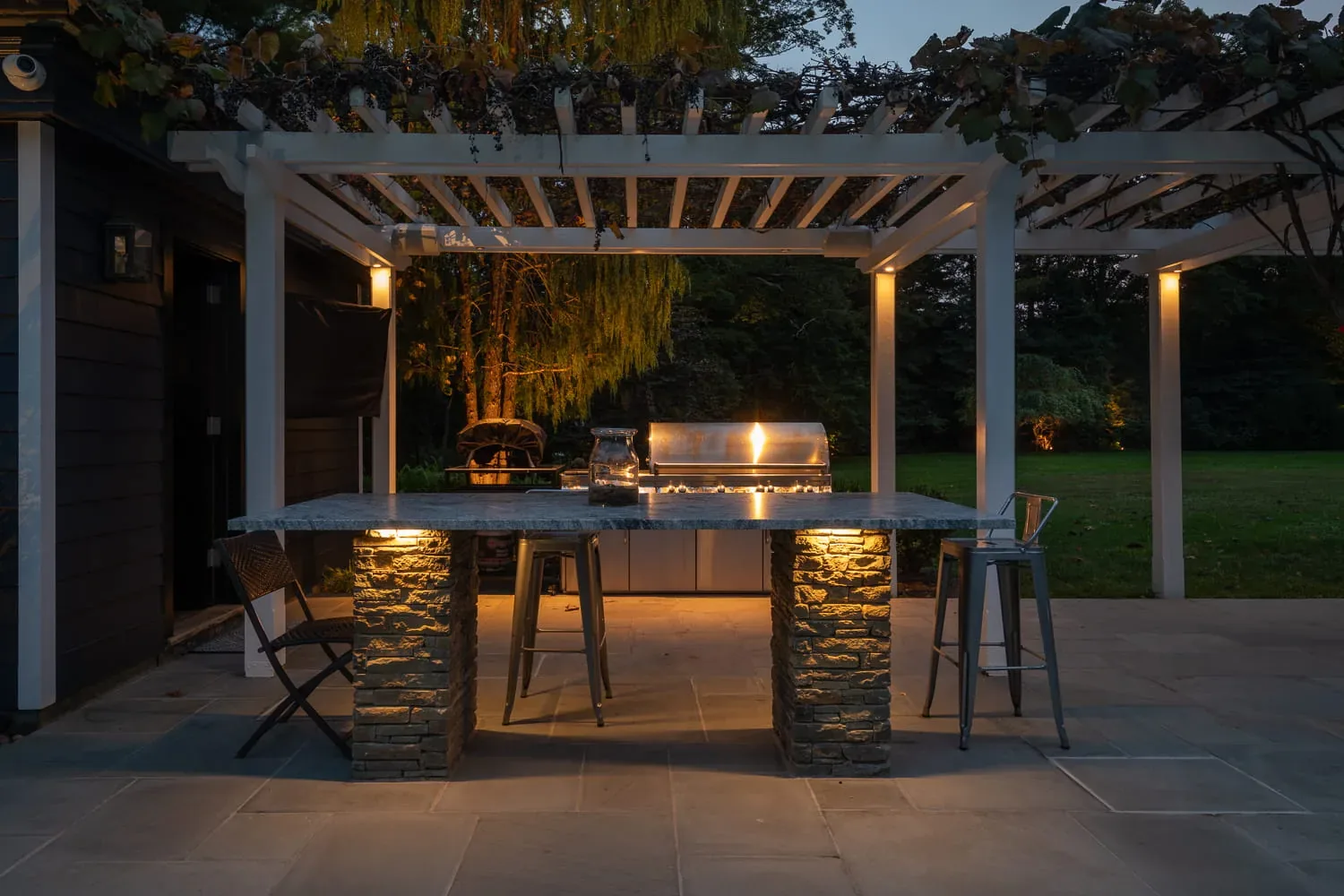 Outdoor kitchen counter and grill lit beneath a white pergola.