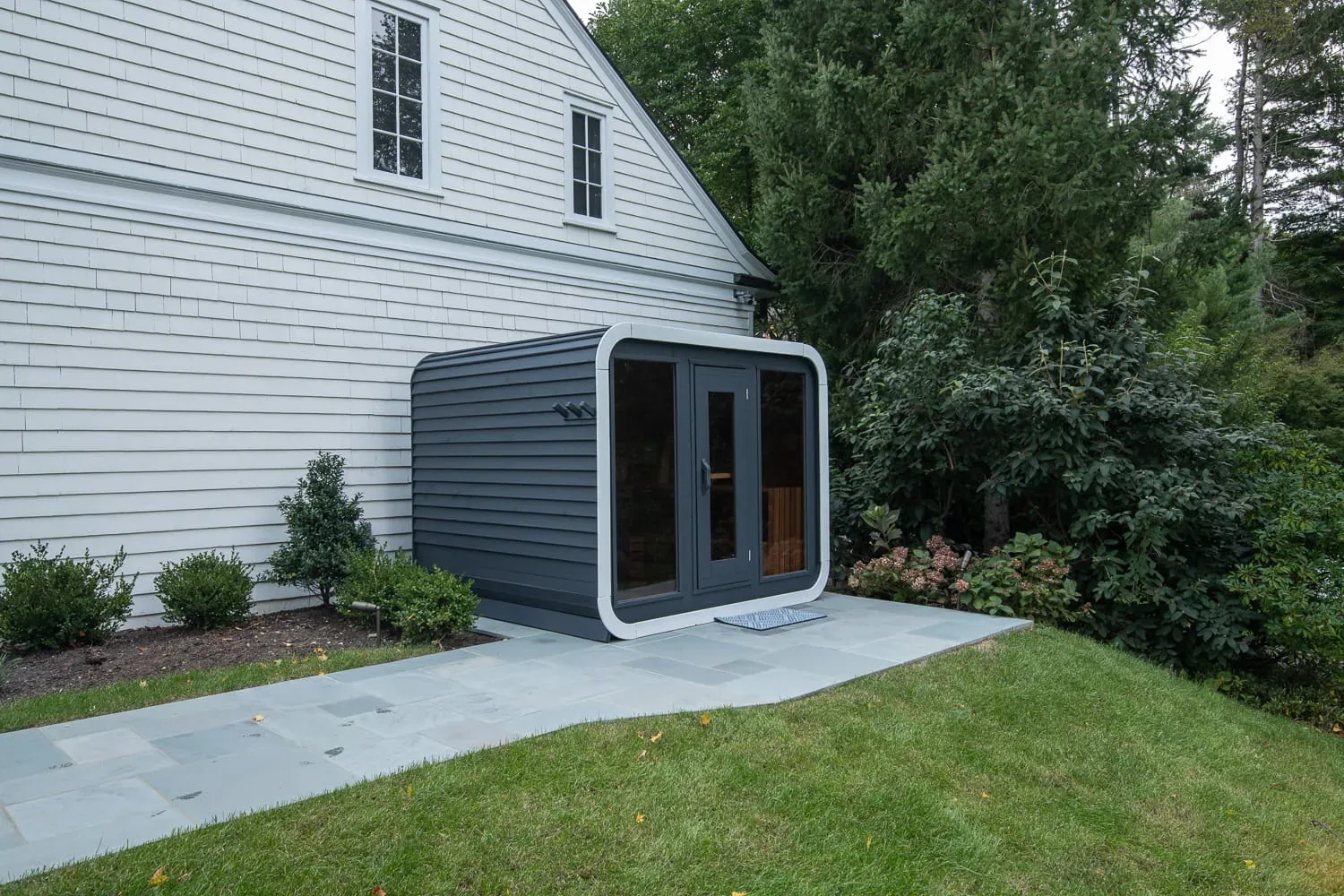 Outdoor sauna beside a white house, lawn, and stone path.