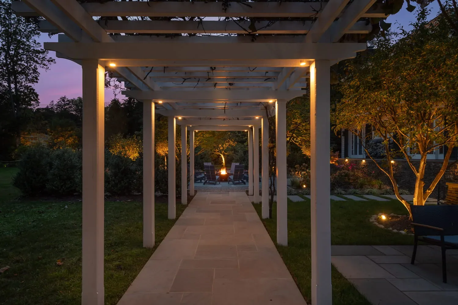 Illuminated pergola walkway leading toward a garden fire pit at dusk.