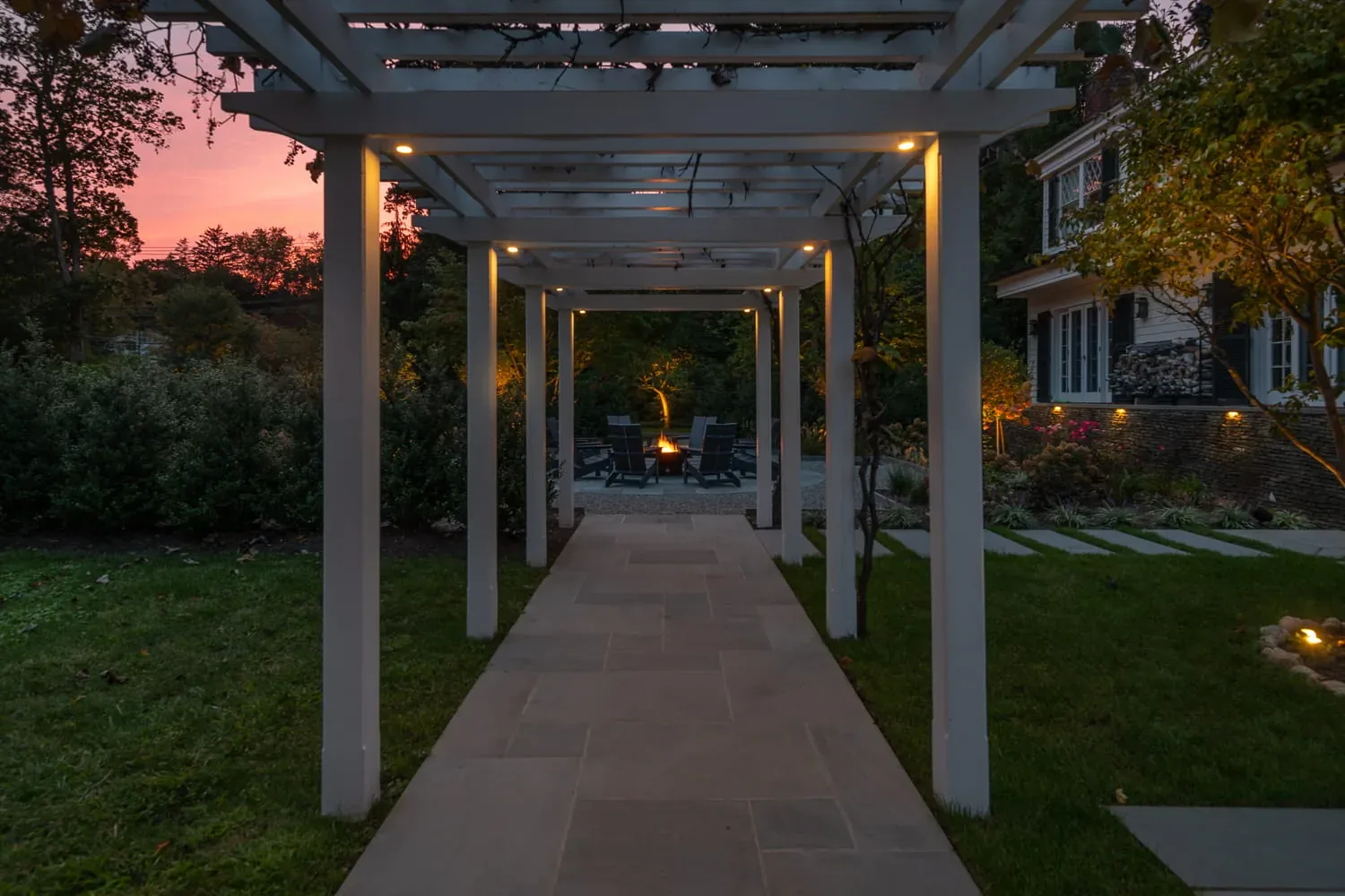 Illuminated pergola walkway leading toward a garden fire pit at dusk.