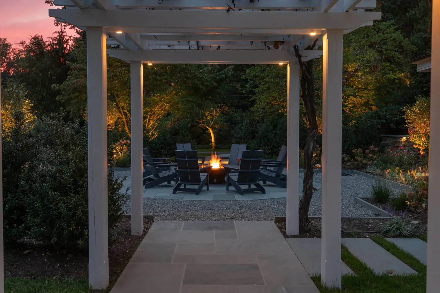 Fire pit and seating viewed through an illuminated white pergola.