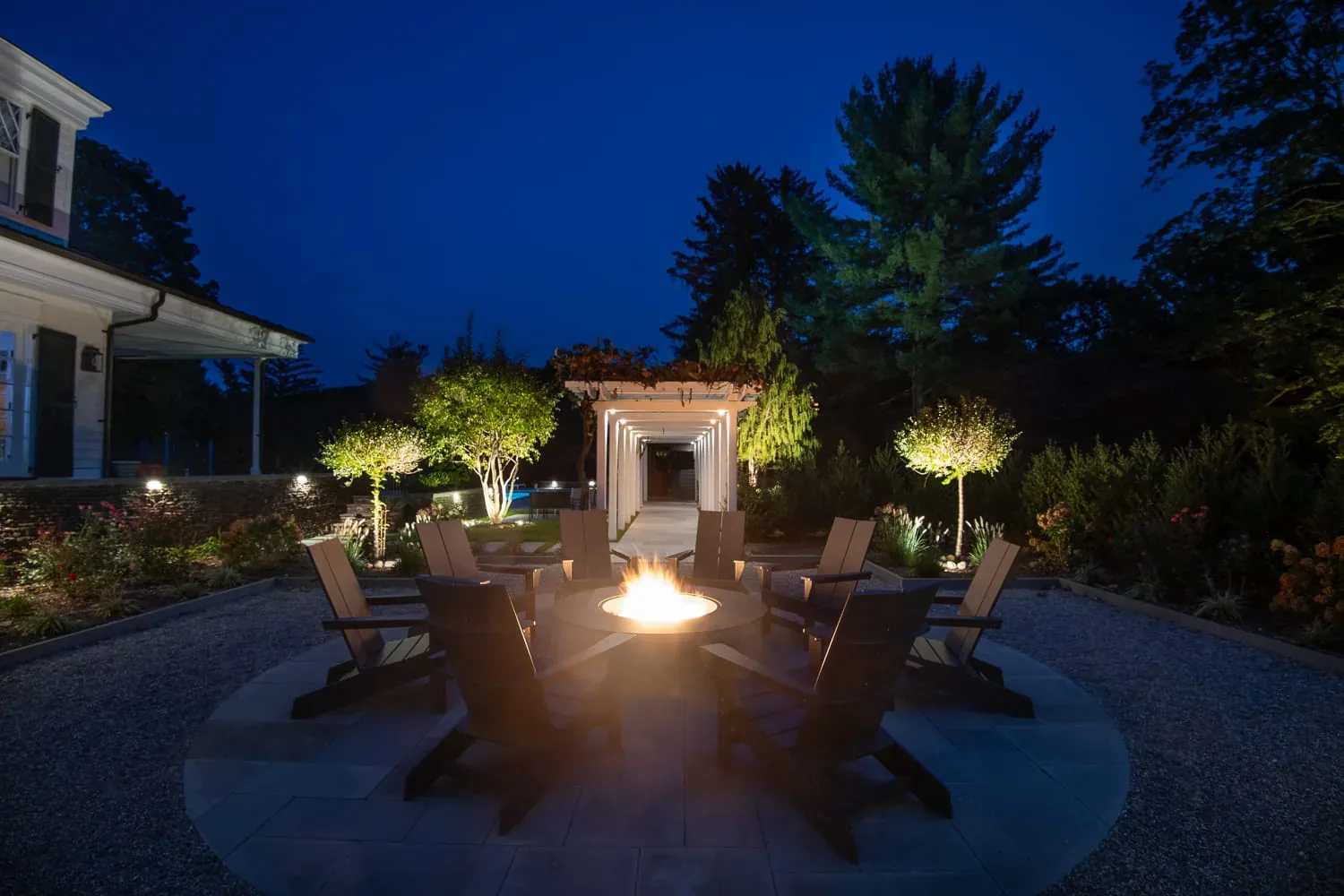 Burning fire pit and chairs surrounded by illuminated garden trees at night.