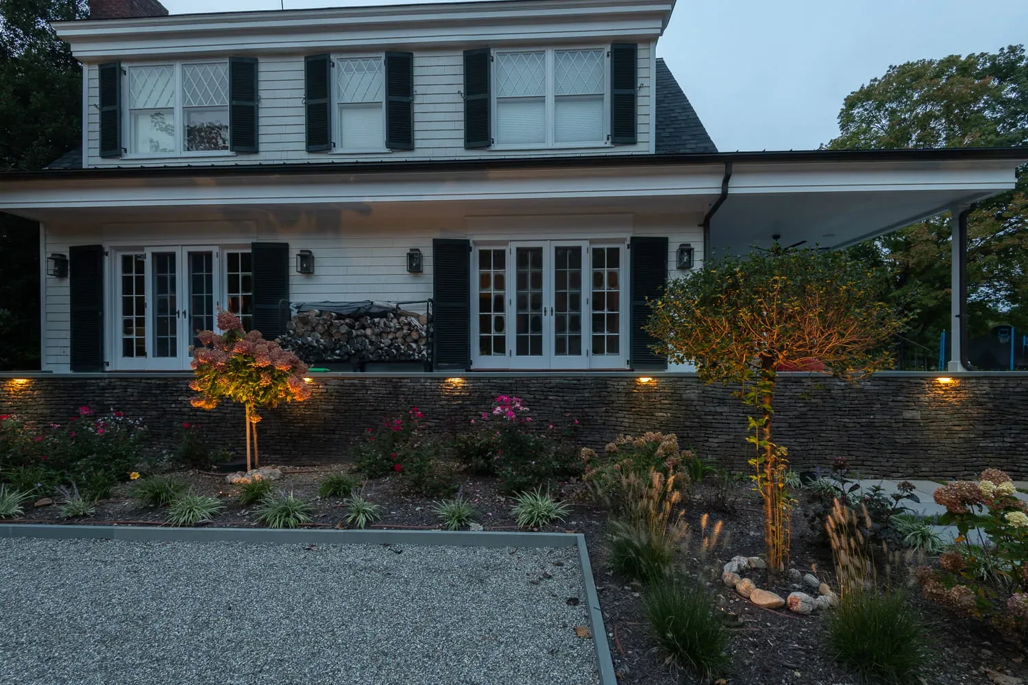 Small trees and planting beds illuminated in front of a farmhouse at dusk.
