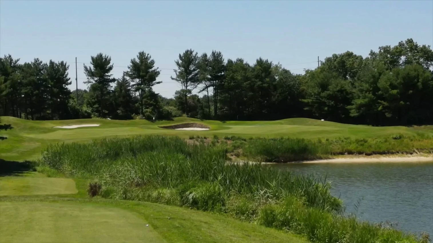 Waterside view across foreground tee and reeds to distant par-three green and pine skyline. Original view of Scotland Run Golf Club.