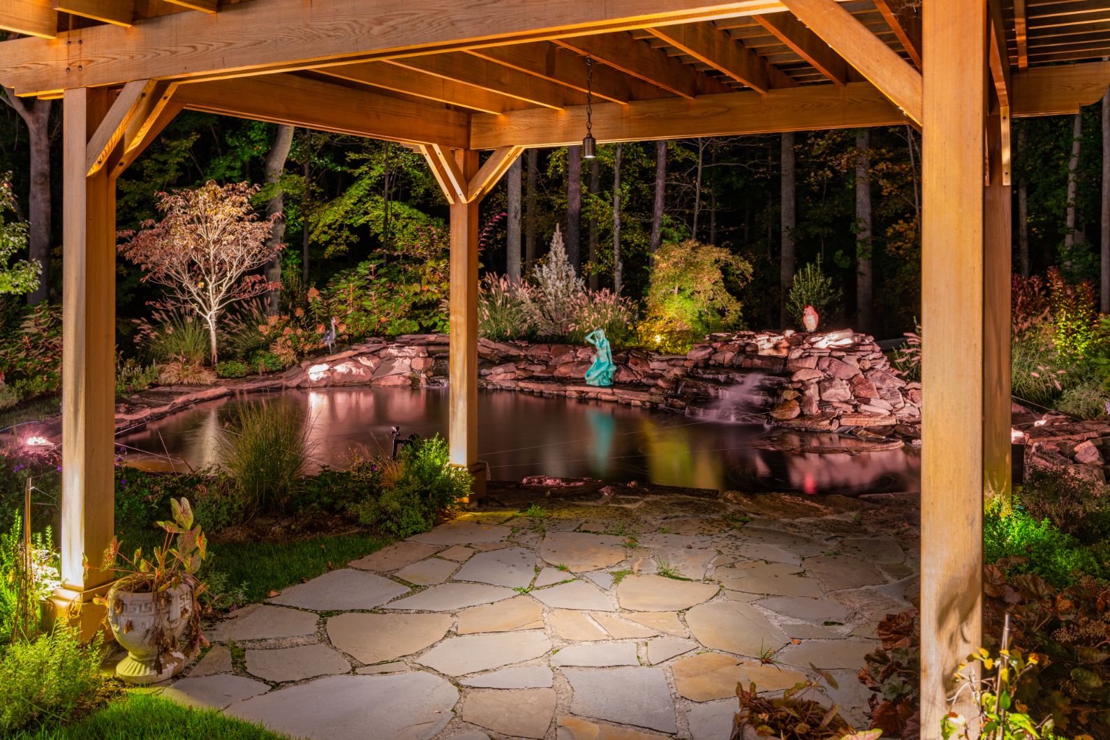 Lighting illuminates a timber pavilion, stone path, pond, and surrounding garden at night.